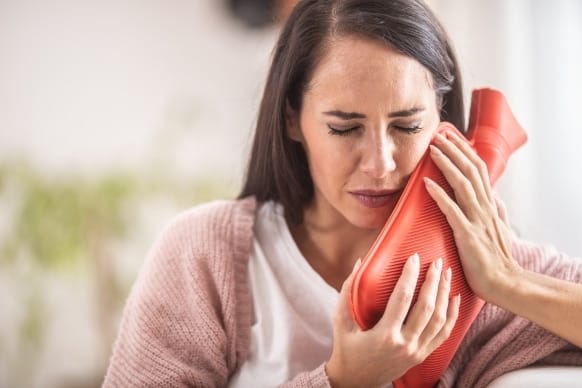 Woman experiencing wisdom tooth pain using a hot water bottle for relief, highlighting methods to alleviate discomfort.