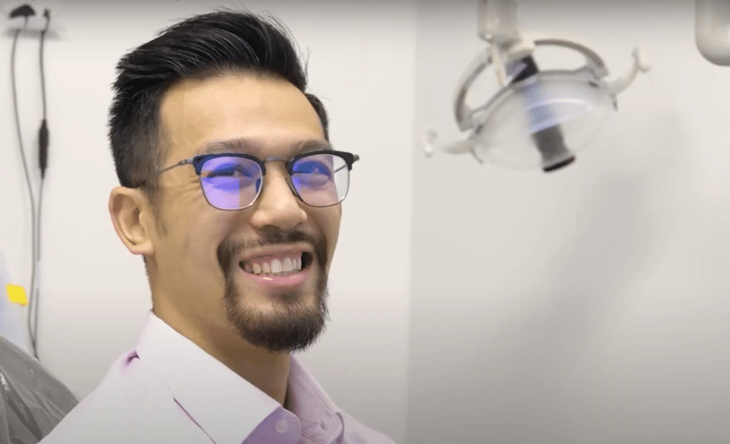 Smiling man with glasses sitting in a dental clinic, representing a successful smile makeover at Smile Avenue Katy.
