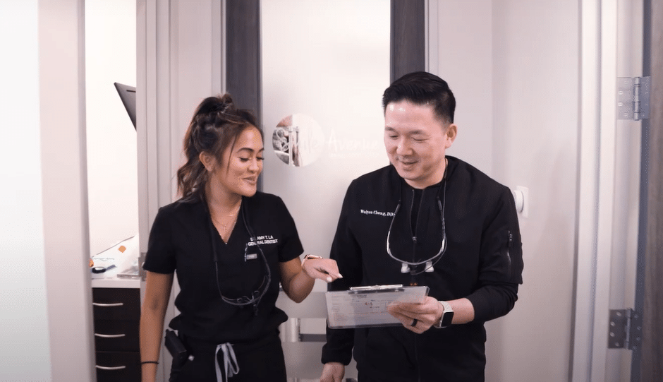 Two dental professionals, a man and a woman, wearing black scrubs and smiling, are engaged in a conversation while holding a clipboard. They are standing in a dental office setting with a partially open door and dental equipment visible in the background. This image is related to patient education and dental care tips, showcasing a friendly and professional environment.