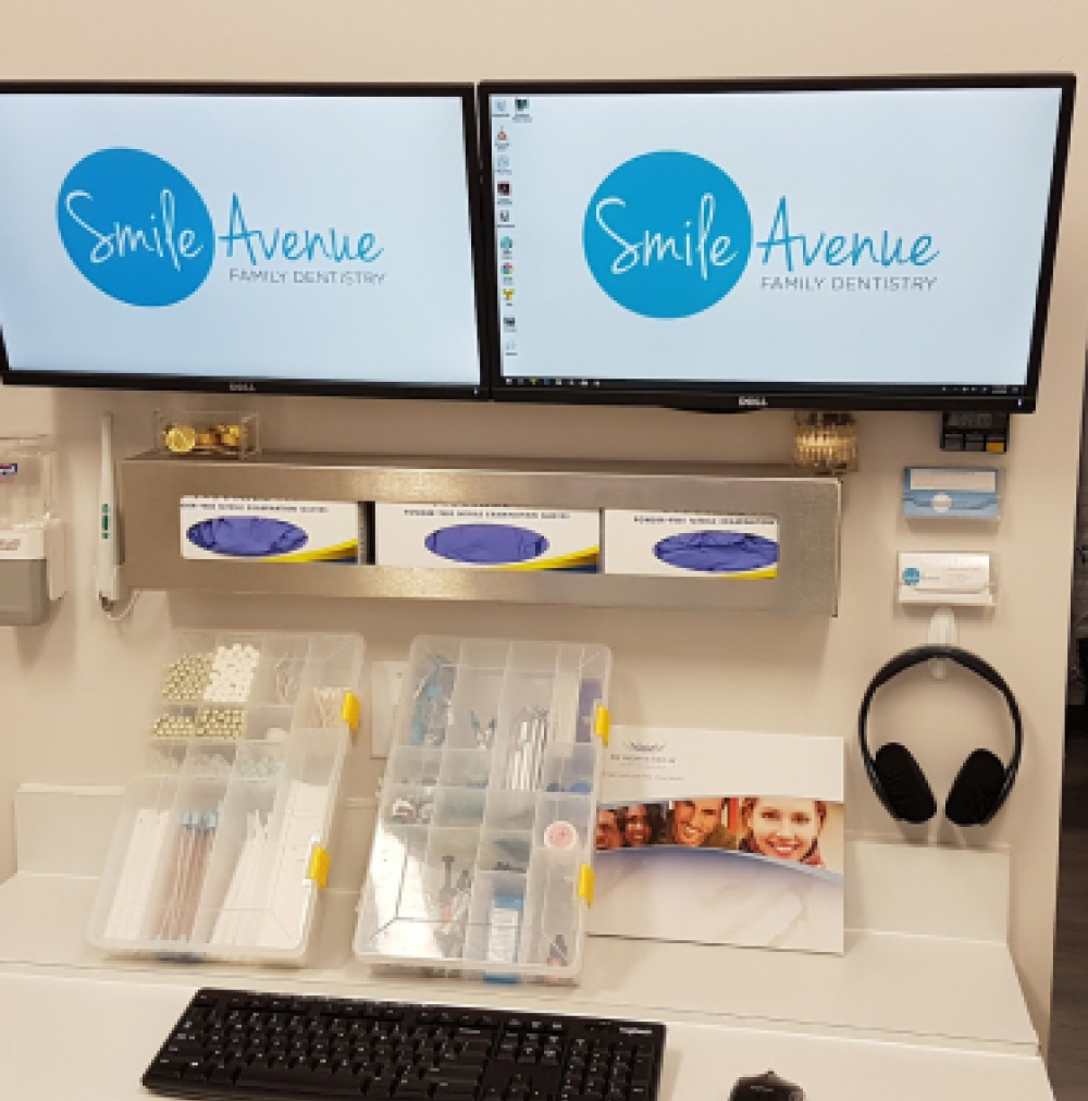 Dental office setup at Smile Avenue Family Dentistry in Cypress, TX, featuring dual monitors displaying the clinic's logo, organized dental tools, gloves, and dental care literature on a countertop, emphasizing comprehensive family and cosmetic dental care.