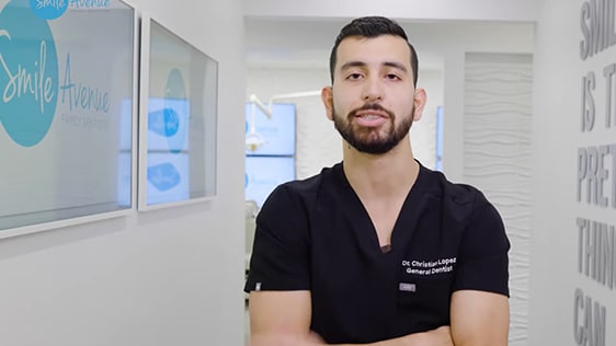 A male dentist in black scrubs standing in a modern dental office with a Smile Avenue sign in the background. Katy Dental Video Library focuses on patient education and smile tips.
