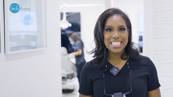 Female dentist smiling in a modern dental clinic, wearing a black uniform with dental equipment in the background. Patient education and smile enhancement tips.