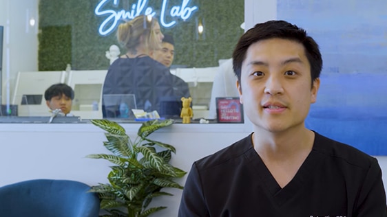 A dentist in a dental office setting, speaking directly to the camera. The background shows a reception area with a neon sign that reads 'Smile Lab.' There are other staff members and patients visible behind a glass partition. A potted plant is in the foreground, emphasizing a welcoming environment. Keywords: dentist, dental office, patient education, smile tips, Smile Lab.
