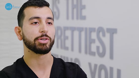 A dentist in a black uniform speaks in a dental office, with blurred motivational text on the wall behind him, as part of Katy Dental's video library focused on patient education and smile tips.