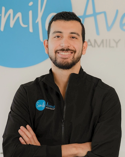 Male dentist in black uniform with Smile Avenue logo, standing confidently with arms crossed in front of Smile Avenue sign at Smile Avenue in Katy.