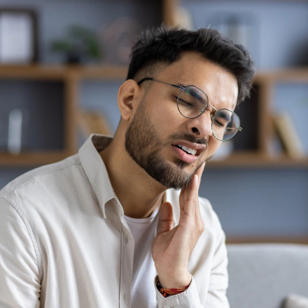 A man in discomfort touching his cheek, possibly experiencing tooth pain, suggesting a need for emergency dental care in Katy, TX. He is wearing glasses and a light-colored shirt, set against a blurred background of shelves.