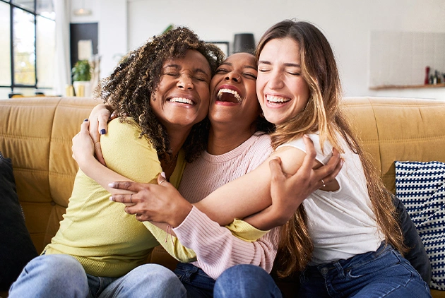 Three joyful women hugging and laughing on a sofa, symbolizing the happiness and comfort experienced after dental check-ups and fillings at Smile Avenue in Cypress, TX.