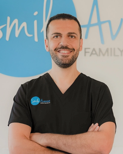 A dentist in a black uniform stands smiling with arms crossed in front of a Smile Avenue logo background.