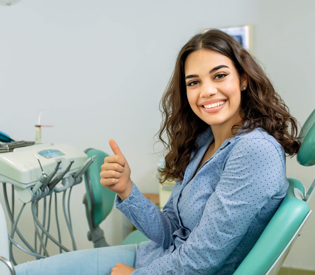 A smiling woman sitting in a dental chair gives a thumbs-up, indicating satisfaction with the oral surgery and family dental care services in Katy, TX. The dental setting includes equipment typical of a professional dentist's office, representing trusted expert care in a friendly environment.