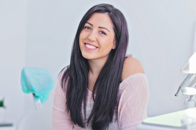 Smiling woman with long dark hair sitting in a dental office chair, showcasing bright white teeth and excellent dental care results at Smile Avenue in Katy, TX.
