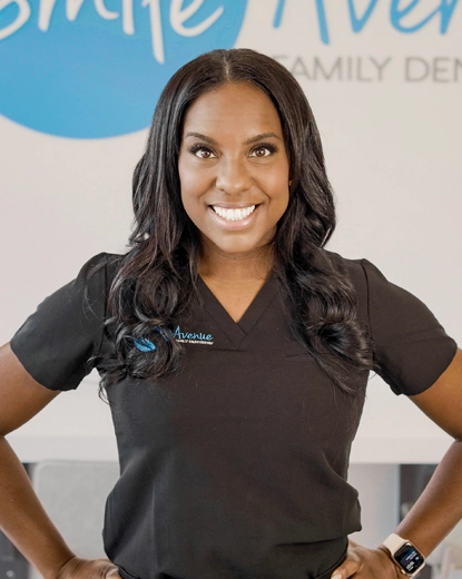 A smiling dentist at Smile Avenue in Katy wearing a black uniform with the clinic's logo. The background features a logo wall of Smile Avenue Family Dentistry.