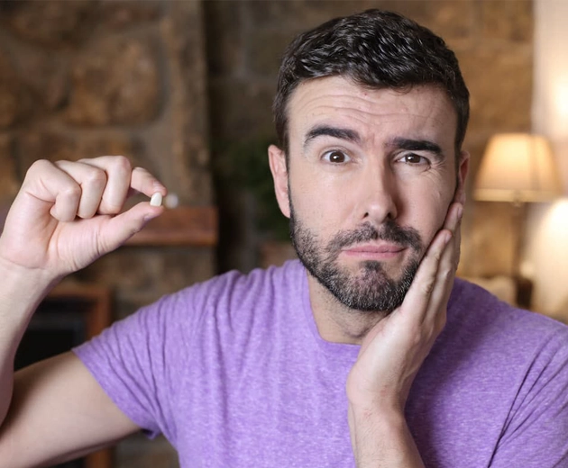 Man in a purple shirt holding his cheek, looking in discomfort, and displaying a small tooth-like object between his fingers, possibly highlighting a tooth extraction or wisdom teeth removal issue in Cypress, TX.