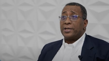 Middle-aged man in a suit speaking, likely sharing his smile makeover experience at Smile Avenue Katy, against a textured white wall backdrop.