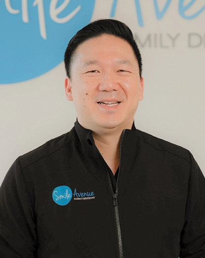 Portrait of a smiling dentist wearing a Smile Avenue Family Dentistry jacket, standing in front of a sign with a blue logo at Smile Avenue in Katy.