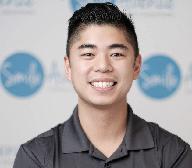Portrait of a smiling dentist in a professional setting, wearing a gray collared shirt with a background featuring Smile Avenue branding. Katy TX Family & Cosmetic Dentist.