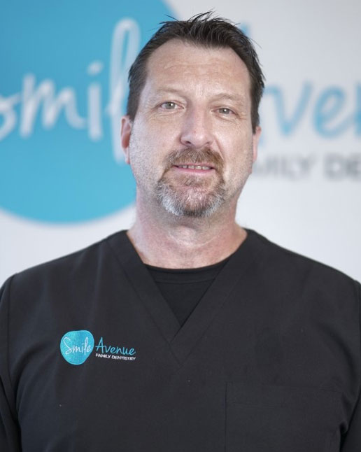 A male dentist in black scrubs stands in front of a sign for Smile Avenue Family Dentistry, representing comprehensive family dentistry services in Cypress and Katy, TX.