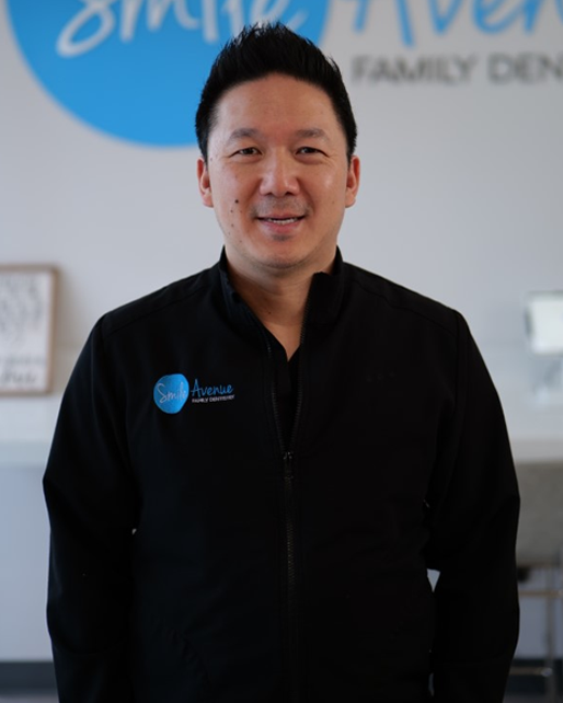 A dentist wearing a black jacket with a blue Smile Avenue logo stands in a dental office. The background features dental equipment and signage. Top Cypress and Katy family dentist and emergency dental care.