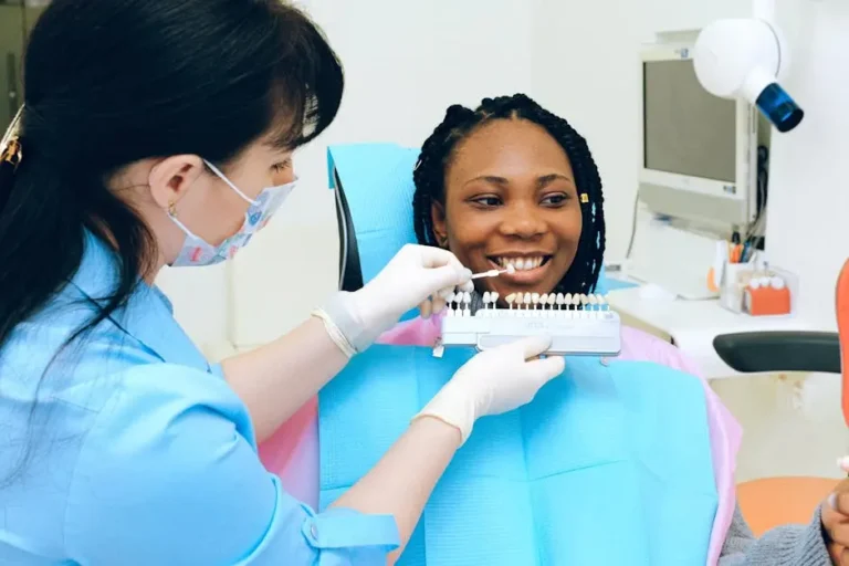 A Female Dentist In A Blue Uniform And Mask Is Holding A Tooth Shade Guide Against A Smiling Woman'S Teeth, Seated In A Dental Chair. This Image Represents Dental Cosmetic Care At Smile Avenue In Cypress, Tx.
