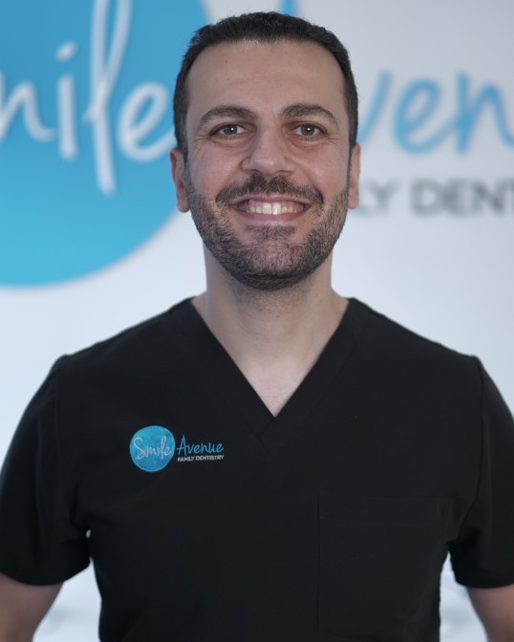 Smiling dentist in black scrubs with Smile Avenue Family Dentistry logo, standing in front of a dental clinic sign. Cypress & Katy family dentist offering emergency dental care.