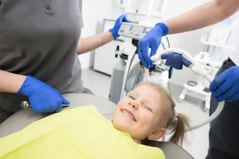A Young Child Smiling And Winking While Sitting In A Dental Chair With A Yellow Bib, Attended By Dental Professionals Wearing Blue Gloves, Highlighting Family-Friendly And Emergency Dental Care In Cypress And Katy.