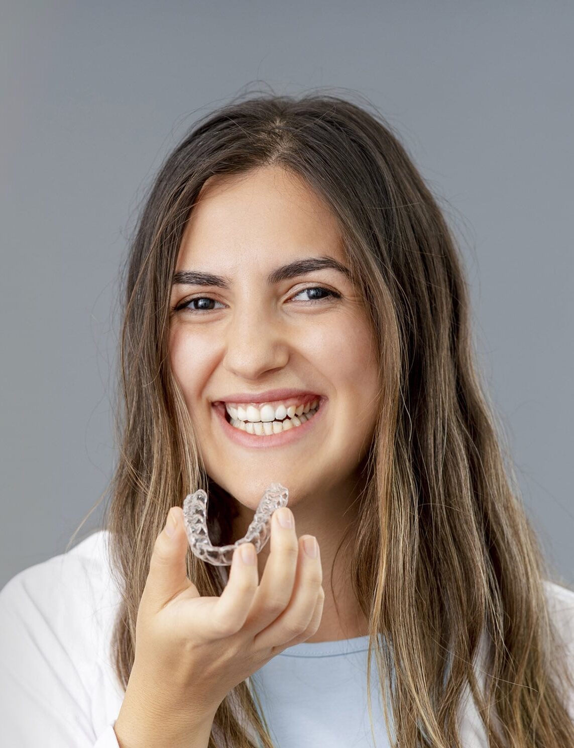 Invisalign Aligners1 4 Young Woman With Long Brown Hair Holding An Invisalign Aligner And Smiling
