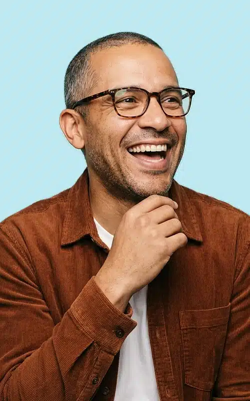 A man in glasses laughing, photographed against a pale aqua background.